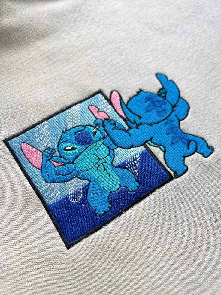 Zwei Stitch-Figuren beim Spielen unter der Dusche, gestickt auf hellgrauem Hoodie
