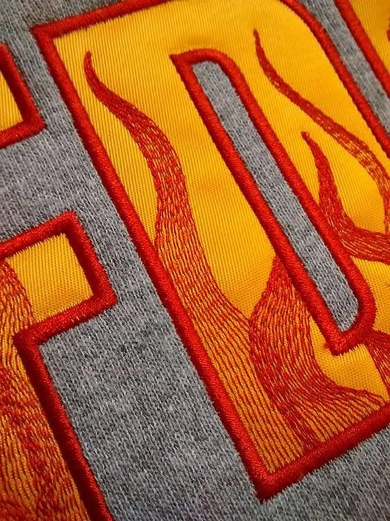 Stickerei mit gelber Applikation und rotem Flammenmotiv auf grauem Sweatshirt, Nahaufnahme